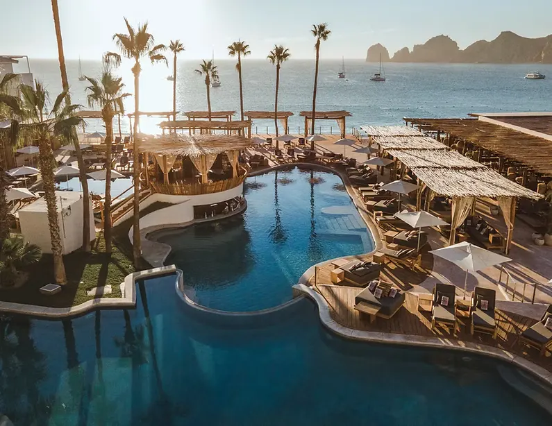 A beach club pool deck under palm trees in Cabo San Lucas, with Land's End across the bay.