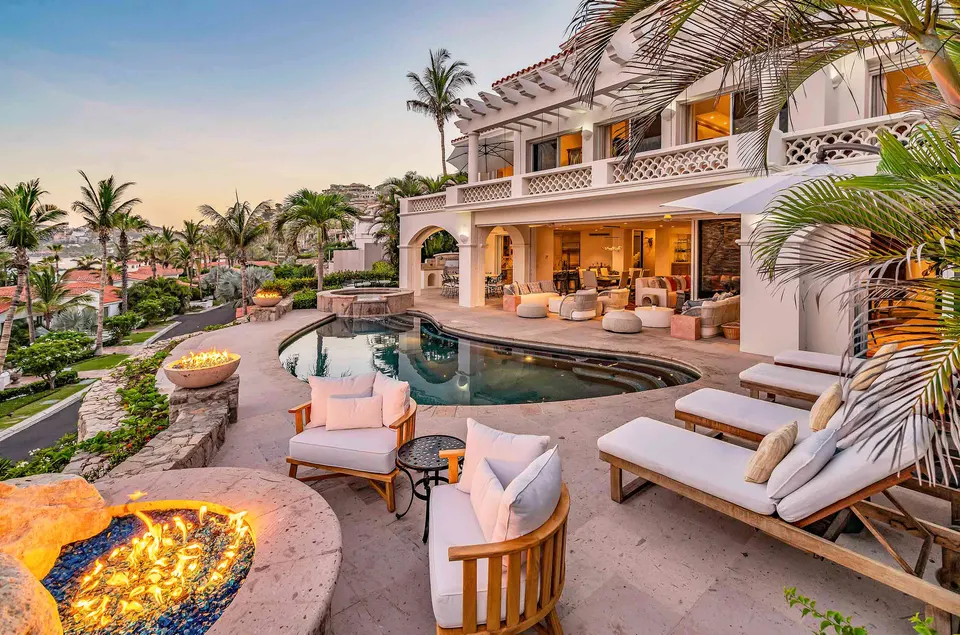 A white hillside villa in Los Cabos at dusk, with a pool, fire pits and loungers under palms.